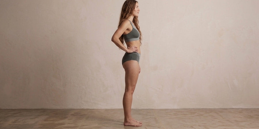 A woman stands in profile wearing a matching gray sports bra and high-waisted athletic brief set against a neutral beige wall and wooden floor.